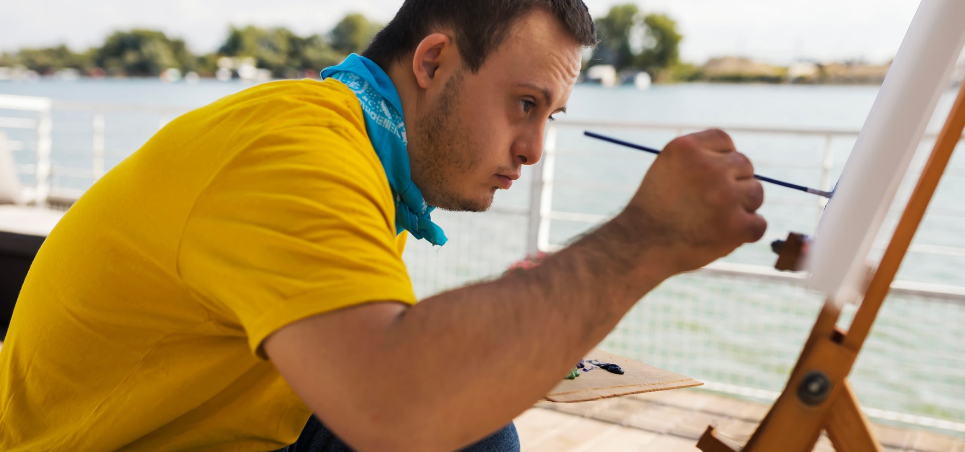 Giovane artista intento a dipingere su una tela, all'aperto vicino a un lago, indossando una maglietta gialla e una bandana azzurra