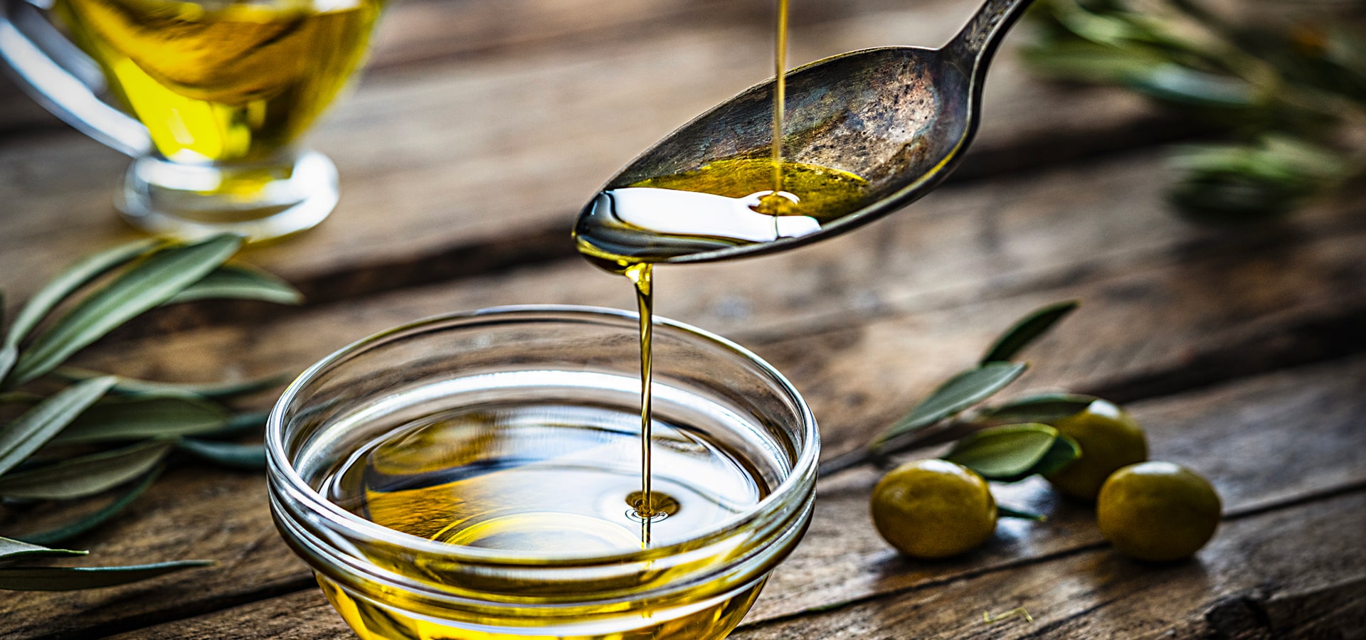 Cucchiaio che versa olio d'oliva in una ciotola di vetro, con olive e foglie di ulivo su un tavolo rustico di legno